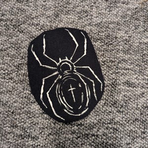 May include: Black patch with a white spider design. The spider has eight legs and a cross in the center of its body. The patch is an oval shape.