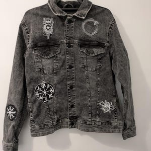 May include: A charcoal gray acid-wash denim jacket featuring a classic collar, button-front closure, and two chest pockets. The jacket is decorated with several white embroidered patches, including a Viking figure, an Ouroboros, and a Vegvisir symbol.