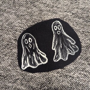 May include: Black fabric patch featuring two white ghost designs. The ghosts are depicted with simple, rounded forms and open eyes and mouths. The patch is set against a textured grey background.