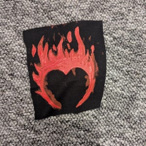 May include: Black fabric patch featuring a red heart design engulfed in flames. The heart shape is outlined in red with a flame-like pattern surrounding it. The patch is likely for clothing or accessories.