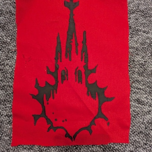 May include: A red fabric patch with a black gothic castle design. The castle silhouette has a dripping effect, with an ornate shape. The patch is rectangular and made of a woven material.
