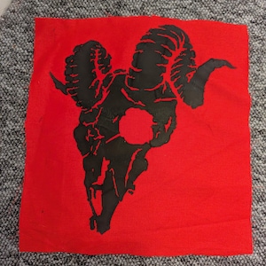 Op de afbeelding: Een rode stoffen vierkant met een zwart ram schedel ontwerp. De ram schedel is weergegeven in een gedurfde, grafische stijl, met gedetailleerde hoorns en een centrale opening. Het ontwerp is gecentreerd op de rode achtergrond.