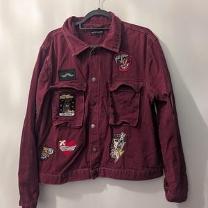 Può includere: Giacca di jeans bordeaux con diverse toppe. Le toppe includono un serpente, una carta dei tarocchi, un cuore, una farfalla e una donna con un fulmine. La giacca ha due tasche sul petto e una chiusura con bottoni.