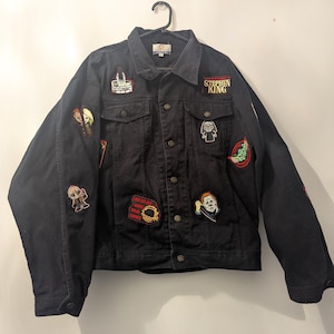 May include: Black denim jacket featuring multiple patches, including a "Stephen King" patch and horror film character patches. The jacket is suspended on a black hanger against a white wall.