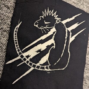 Może przedstawiać: Czarna naszywka z tkaniny z białą ilustracją szczura z koroną i zakrzywionym ogonem. Białe ukośne linie znajdują się za szczurem.