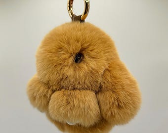 May include: A fluffy, tan-colored rabbit keychain with a gold-colored metal ring and a brown leather strap. The keychain is made of soft, plush material, and the rabbit has a small, dark eye.