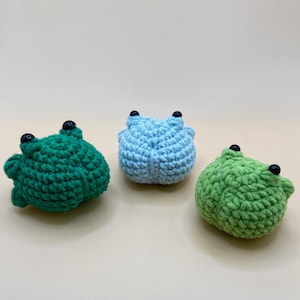 Pode incluir: Três brinquedos de crochê em forma de sapo em tons de verde e azul. Cada sapo tem olhos de botão pretos e um corpo arredondado. Os sapos são feitos à mão e parecem macios e felpudos.