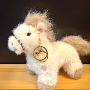 May include: A plush white pony with a tan mane and tail. The pony has a brown bridle and a gold-colored keychain with a horseshoe design. The pony is standing on a wooden surface against a dark background.