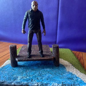 Può includere: Una action figure di Jason Voorhees è in piedi su un molo di legno in miniatura sopra una base d'acqua blu. La figura indossa una maschera da hockey, una giacca nera e pantaloni scuri. Il molo è affiancato da due pali di legno.