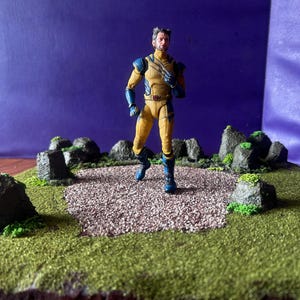 Può includere: Una action figure di Wolverine in costume giallo e blu è su un diorama in miniatura. Il diorama presenta un sentiero di ghiaia circondato da erba verde e rocce grigio scuro, su uno sfondo viola.
