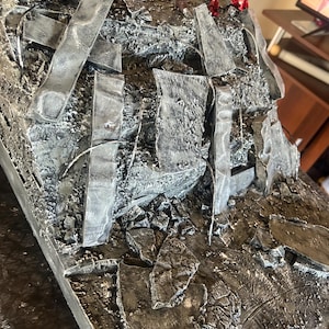 Può includere: Una base diorama dettagliata con una combinazione di colori strutturati, grigi e argentati. La base presenta strutture frammentate dall'aspetto metallico e detriti, creando un aspetto logoro dalla battaglia. La base è progettata per esporre action figure.