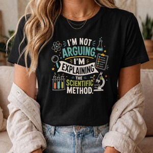 May include: A black short-sleeved t-shirt featuring a colorful graphic with the text I'M NOT ARGUING, I'M EXPLAINING THE SCIENTIFIC METHOD. The design includes illustrations of an atom, a magnifying glass, a beaker, a clipboard, test tubes, and a microscope.