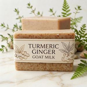 Puede incluir: Dos barras rectangulares de jabón de color marrón claro con motas visibles. La barra frontal tiene una etiqueta que dice "TURMERIC GINGER GOAT MILK" con ilustraciones de jengibre y hojas. Hay vegetación y pequeñas piedras en el fondo.