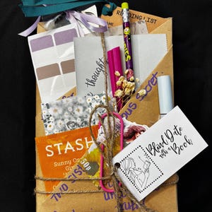 May include: A brown paper package tied with twine, teal and lavender ribbons. Contents include stickers, a pencil, a pink highlighter, a tag that says "Blind Date with a Book", and a tea bag.