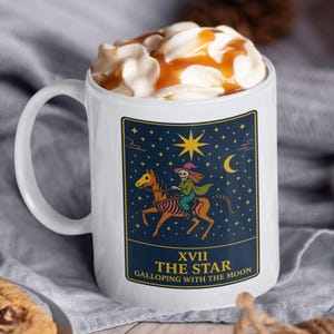 May include: White ceramic mug featuring a tarot card design with a star and moon theme. The mug is filled with whipped cream and caramel sauce. A plate of chocolate chip cookies and cinnamon sticks are also visible.