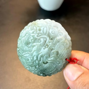 May include: A circular jade pendant featuring a detailed dragon carving. The pendant is predominantly light green with white highlights. A red string is attached. The dragon design is the focal point.