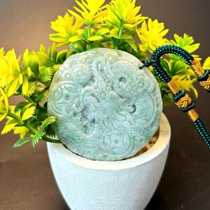 May include: A circular, pale green jade pendant featuring detailed carvings of a mythical beast. The pendant is strung on a dark green and gold cord. It's displayed in a small white pot with yellow flowers.