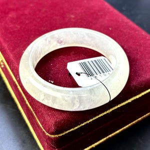 May include: A translucent, off-white bangle bracelet. The bracelet is circular and appears to be made of a stone-like material. A white tag with black text and a barcode is attached. The bracelet rests on a red velvet surface.