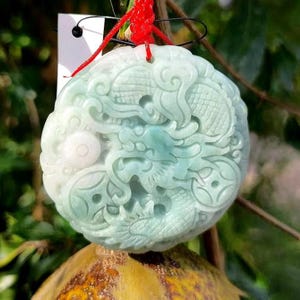 May include: A round, pale green jade pendant with intricate carvings of dragons and swirling patterns. The pendant is suspended by a red cord and a white tag. The jade has a translucent quality, with variations in color.