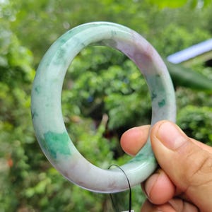 Könnte beinhalten: Ein Jade-Armreif mit glatter, polierter Oberfläche. Das Armband weist eine Mischung aus grünen, weißen und violetten Farbtönen auf. Der Jade ist durchscheinend und lässt Licht durch. Das Armband hat eine kreisförmige Form und ist zum Tragen am Handgelenk bestimmt.