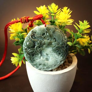 May include: A jade pendant with a carved dragon design, in shades of green and grey, hangs from a red cord. The pendant is displayed in a white pot with yellow flowers and green leaves.