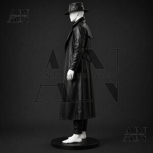 K&ouml;nnte beinhalten: Ein schwarzer Ledertrenchcoat an einer wei&szlig;en Schaufensterpuppe. Die Puppe tr&auml;gt zudem einen schwarzen Fedora-Hut und eine schwarze Hose. Das Ensemble steht auf einem runden schwarzen Sockel. Der Text Antiques Nest ist im Hintergrund zu sehen.