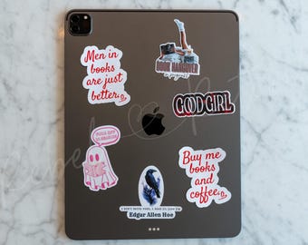 May include: A collection of stickers on a gray tablet. The stickers feature phrases like "Men in books are just better," "Book hangover in progress," and "Buy me books and coffee." Other stickers include a ghost, a raven, and the words "GOOD GIRL."