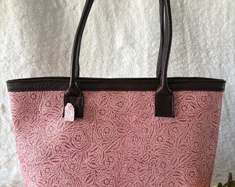 May include: Pink tote bag with a floral embossed design. The bag has dark brown handles and trim. A small tag is attached to the bag. A small cowboy hat and a sunflower are in the foreground.