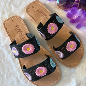 Op de afbeelding: Een paar zwarte leren sandalen met geborduurde bloemen in roze, blauw en paars. De sandalen hebben twee bandjes en de merknaam "Zamarla" is op de bandjes gedrukt. De sandalen liggen op een wit oppervlak.