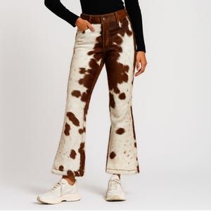 Puede incluir: Pantalones acampanados con estampado de piel de vaca en marrón y blanco. Los pantalones tienen una cinturilla y bolsillos marrones. El conjunto se completa con zapatillas blancas. El estampado de piel de vaca es único.