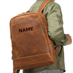 以下が含まれることがあります： 前面に「NAME」とプリントされた茶色の革製バックパック。 バックパックは長方形で、ジッパー付きのフロントポケットと調節可能なショルダーストラップが付いています。 白いステッチがエッジを強調しています。
