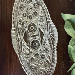 May include: An oval clear glass dish featuring an intricate pressed pattern of geometric shapes, including stars, hexagons, and diamonds. The rim has a scalloped, serrated edge, and the surface displays a complex, multifaceted design throughout.