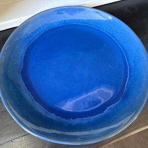 May include: A round ceramic dinner plate featuring a vibrant cobalt blue center that transitions into a lighter, gradient blue rim. The surface has a smooth, glossy finish with subtle variations in color intensity across the circular plate.