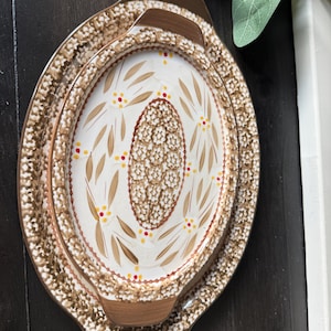 May include: Two oval ceramic serving platters with handles, featuring a cream-colored center with tan leaf motifs, small red and yellow dots, and a central oval pattern of white flowers. The rims have a brown and white floral border.