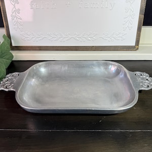 May include: A rectangular silver-toned metal tray with a smooth, shallow basin and ornate, scroll-patterned handles on each side. The tray features a polished, metallic finish and a wide, flat rim, providing a simple yet detailed serving piece.