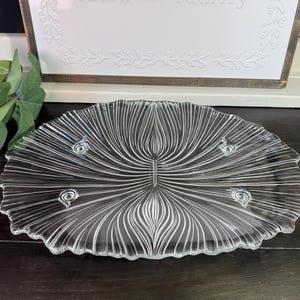 May include: A clear glass serving platter featuring a radial, fan-like embossed pattern radiating from the center. The circular plate has a scalloped edge and is supported by three small, integrated glass feet on the underside.