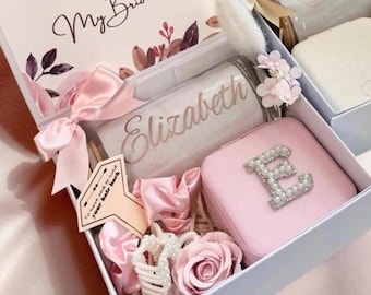 May include: A white gift box containing a card with the text Will You Be My Bridesmaid, a tumbler personalized with the name Elizabeth, a pink jewelry case with a pearl-encrusted letter E, a pink scrunchie, a white hair clip, and a faux rose.