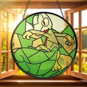Pode incluir: Vitral redondo com uma rã de desenho animado em tons de verde, amarelo e vermelho. A rã está sentada numa folha. A obra de arte é emoldurada em preto e pendurada por uma corrente. A janela está em frente a uma janela aberta com luz solar.