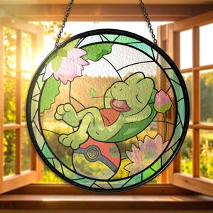Pode incluir: Janela redonda de vitral com um personagem de desenho animado de tartaruga descansando em uma bola vermelha e branca. A tartaruga é verde com uma concha vermelha. Flores de lótus rosa e folhas verdes cercam a tartaruga. A janela está pendurada.