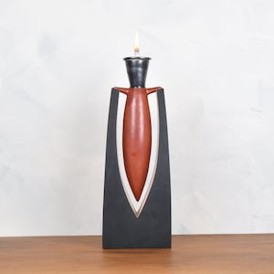 May include: A modern candle holder with a black stand and a red ceramic candle. The candle is lit and the flame is visible.