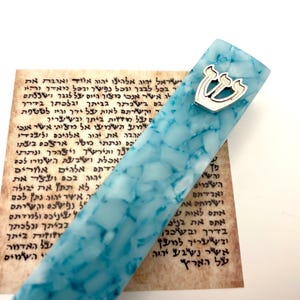 May include: A blue and white mezuzah case with a silver-colored emblem. The case is next to a piece of paper with Hebrew text. The mezuzah case has a marbled appearance.