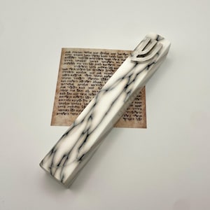 Puede incluir: Un estuche de mezuzá rectangular blanco y negro veteado, coronado por un símbolo Shin plateado. Descansa sobre una superficie blanca junto a un pergamino con texto hebreo.
