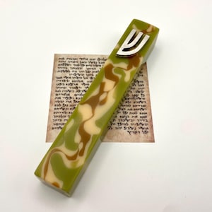 Puede incluir: Una mezuzá decorativa con un patrón veteado verde, marrón y crema. Una letra Shin de color plateado está fijada en la parte superior. La mezuzá descansa sobre un trozo de pergamino con texto hebreo.