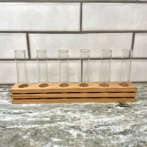 Puede incluir: Un soporte de madera para tubos de ensayo con seis tubos de vidrio transparente. El soporte es de madera marrón claro con un diseño en capas. Los tubos son vacíos y cilíndricos, ideales para plantas.