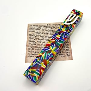 May include: A colorful mezuzah case with a vibrant mosaic pattern of red, yellow, green, and blue. The case has a silver-colored Shin symbol on top. A piece of parchment with Hebrew text is visible in the background.