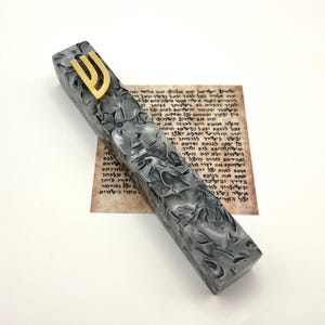 May include: A gray and white mezuzah with a gold-colored Shin symbol on top. The mezuzah has a textured pattern and rests on a paper with Hebrew text. The background is white.