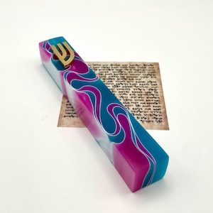 Puede incluir: Un estuche de mezuzá colorido con una letra hebrea Shin dorada. El estuche presenta un diseño vibrante con tonos de azul, rosa y blanco. Un pergamino con texto hebreo es visible en el fondo.