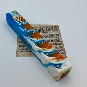 Puede incluir: Un estuche de mezuzá decorativo con un diseño veteado azul, blanco y naranja. El estuche tiene un emblema dorado en la parte superior. Un papel con texto hebreo es visible en el fondo.