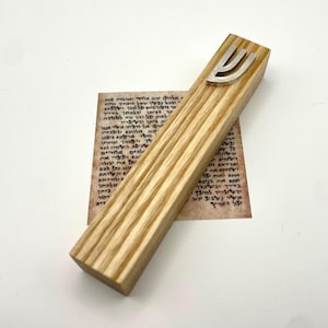May include: A wooden mezuzah case with a silver-colored Shin symbol on top. The case is rectangular with a light wood grain pattern. A small piece of parchment with Hebrew text is visible beneath the case.