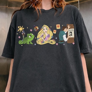 Può includere: T-shirt grigio scuro con una grafica colorata di personaggi delle fiabe. Il design include un sole, un camaleonte verde, una principessa bionda e un uomo con un poster "Wanted". La maglietta è realizzata in materiale morbido.